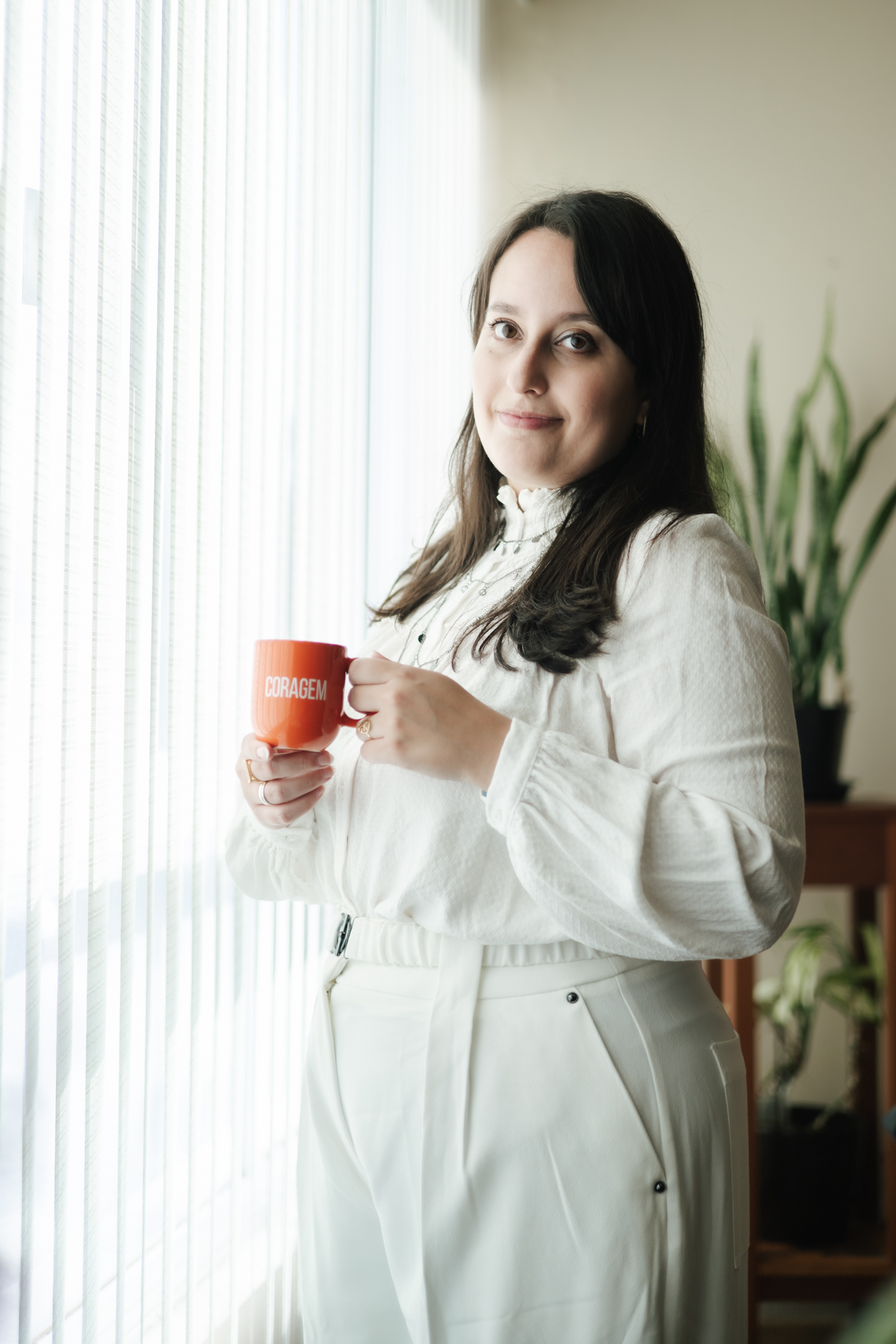 Amanda sorrindo com uma caneca nas mãos, ao lado de uma janela iluminada